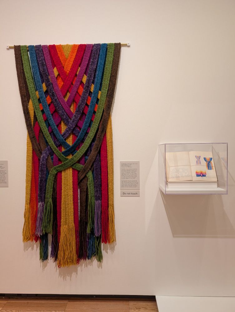 A photograph of an installation showing a large hand-woven tapestry with jewel tones of orange, red, purple, blue, olive, and brown hanging on a white-painted wall adjacent to a vitrine showing an artist's sketchbook