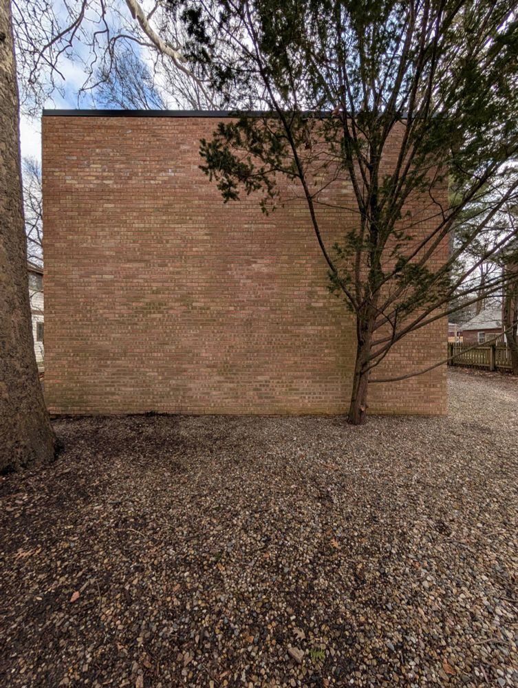 Photograph of the front face of a house showing a two story brick wall with no windows, a yard of river stones, and a small cedar tree