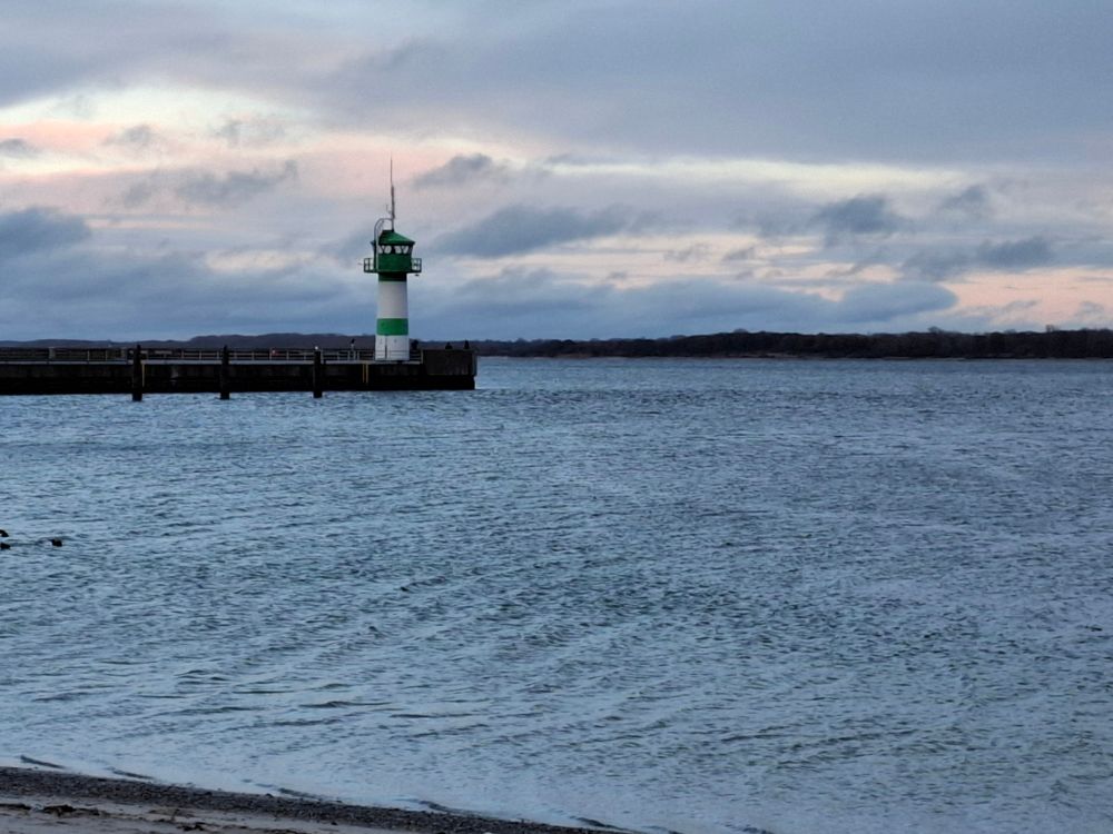 Meer. Im unteren Bereich etwas Strandanschnitt. Ein grün,weißer Leuchtturm auf einer Seebrücke. Der Himmel ist wolkig