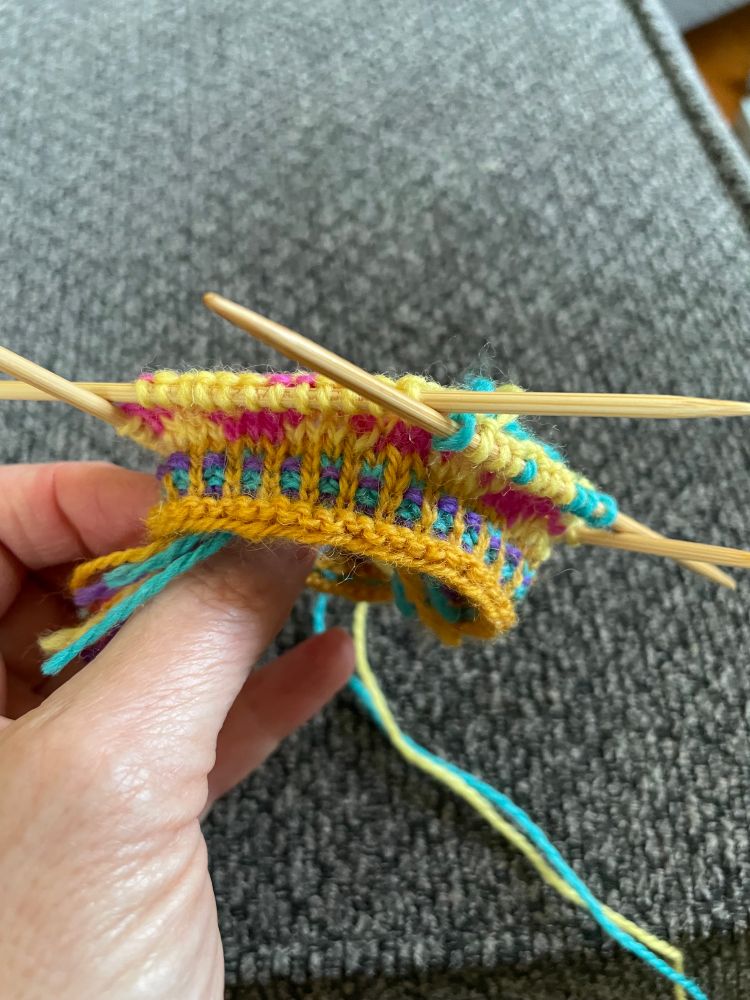 Hand holding bright yellow, pink & aqua colored fair isle cuff on double pointed knitting needles.
