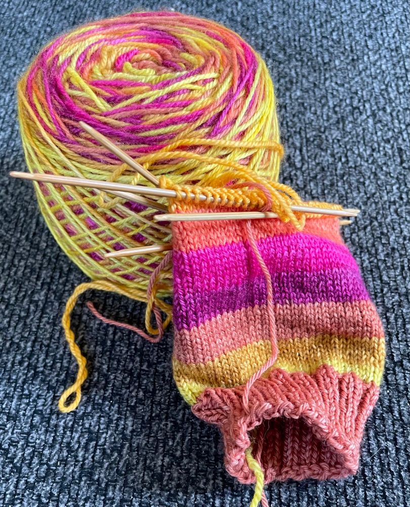 A brightly colored sock with stripes in shades of yellow, orange, and magenta being knit on double-pointed needles