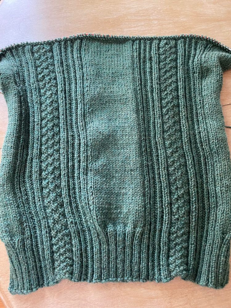 A green handknit sweater with ribs & textured stitches on a long circular knitting needle.