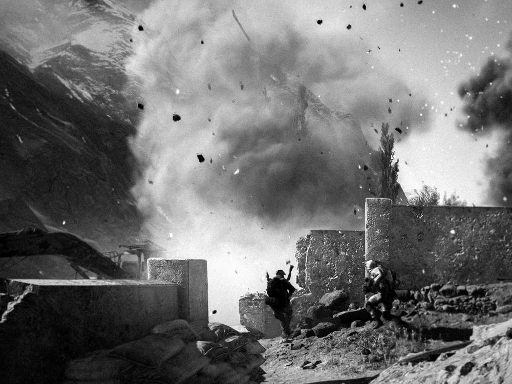 A photograph of a dust cloud and debris from an explosion behind a damaged wall on the Liberation Peak map. A soldier armed with a RPG stands in front of the cloud and another soldier is rushing towards them.