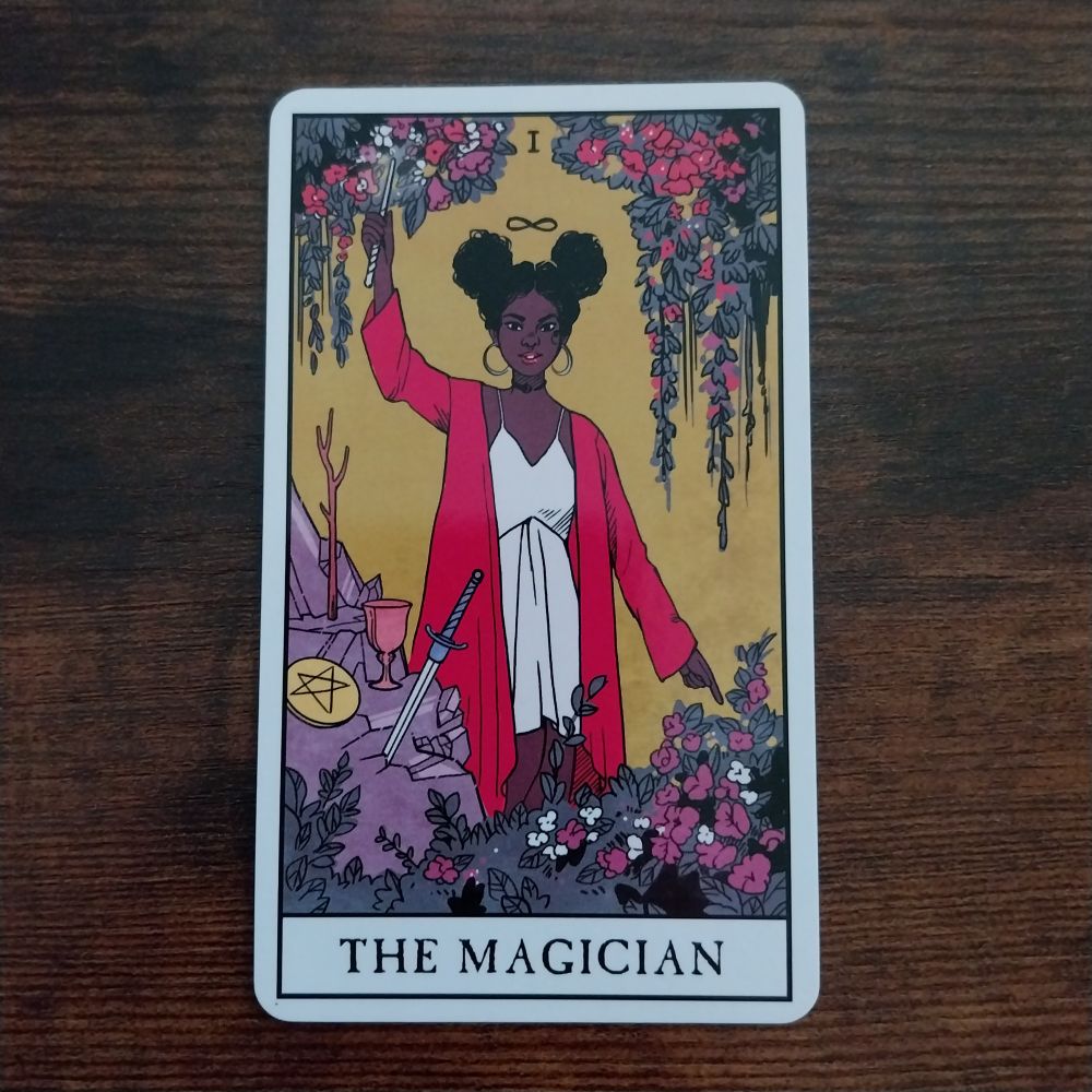 I The Magician Tarot card from the Modern Witch Tarot by @lisasterle.bsky.social depicting a Black woman as The Magician, dressed in red and white with an infinity sign above her head and pointing a wand upwards with her right hand and her left hand downwards, connecting heaven and earth. In front of her are representations of the four Tarot suits and the background is yellow and bordered by roses and lilies
