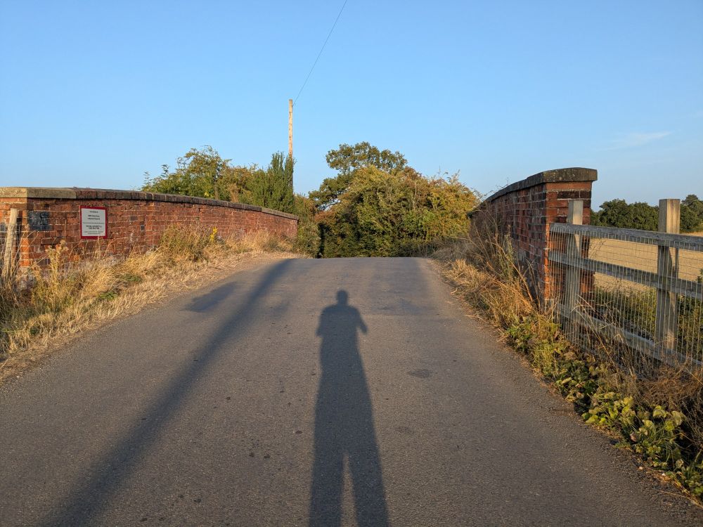 A rural lane, loping over a bridge. Two shadows, of a pole and of the photographer, are on the tarmac.