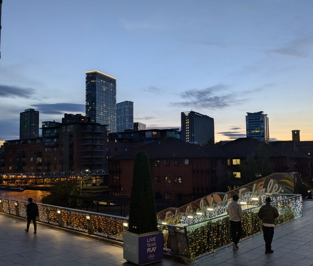 A city landscape, shortly after sunset. The sky is almost fully clear. In the background are several high rise buildings. Centre left shows part of a canal. In the foreground are walkways with various subtle lighting.