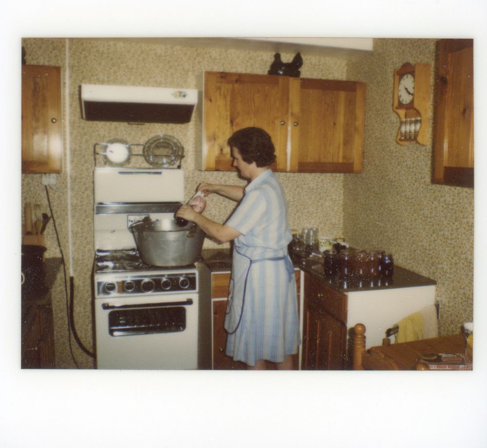 My mom making jams in the kitchen of our house. There are several varieties on the cupboard surface, in jars. These are some combination of blackberry, raspberry, marmalade, strawberry, various plum jams, damson, greengage, loganberry, and rhubarb and ginger. Jam is being collected from a large stove pan with a gravy boat. There are wall cupboards, and a kitchen clock.