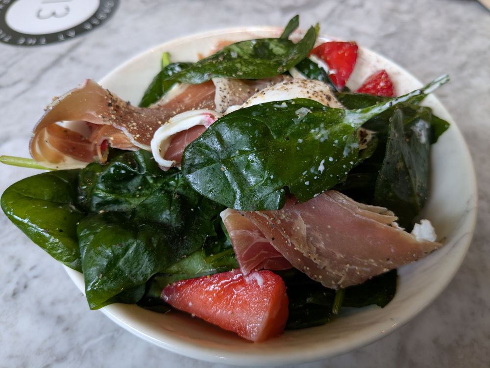 A small bowl of salad leaves, strawberries, burrito and thin ham, on a table.