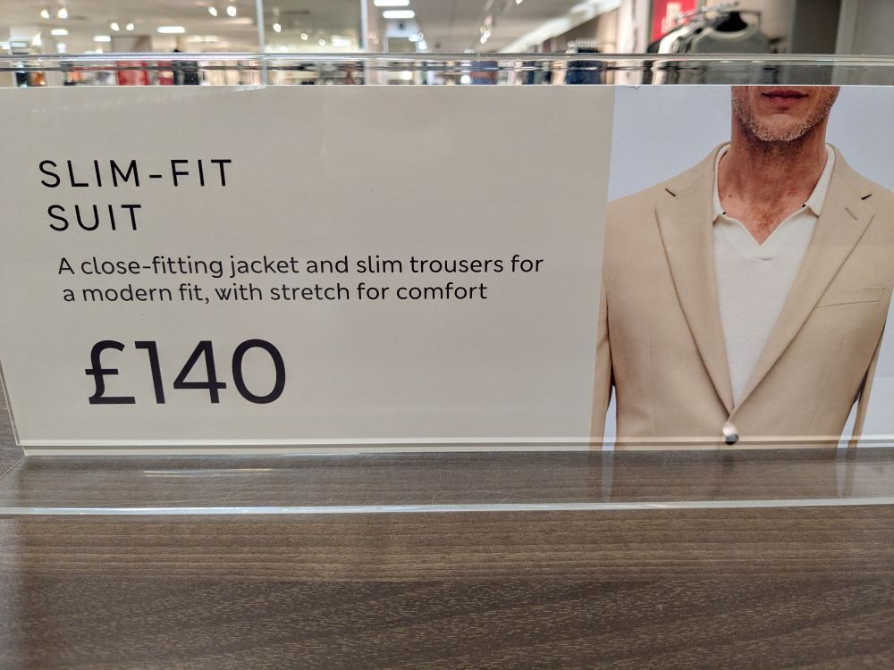 The sign on a display in a branch of Marks and Spencer in Worcester, England. It says "SLIM-FIT SUIT A close-fitting jacket and slim trousers for a modern fit, with stretch for comfort. £140" Next to the text is a picture of the upper torso (and chiselled jawline) of a man wearing a cream-coloured jacket over a white top.