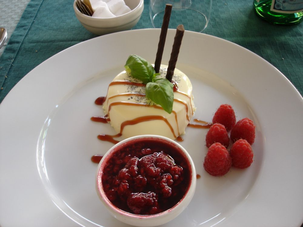 A small, individual, panna cotta, on a large dessert plate. Some raspberries are nearby, as is a small pot of pureed fruit.