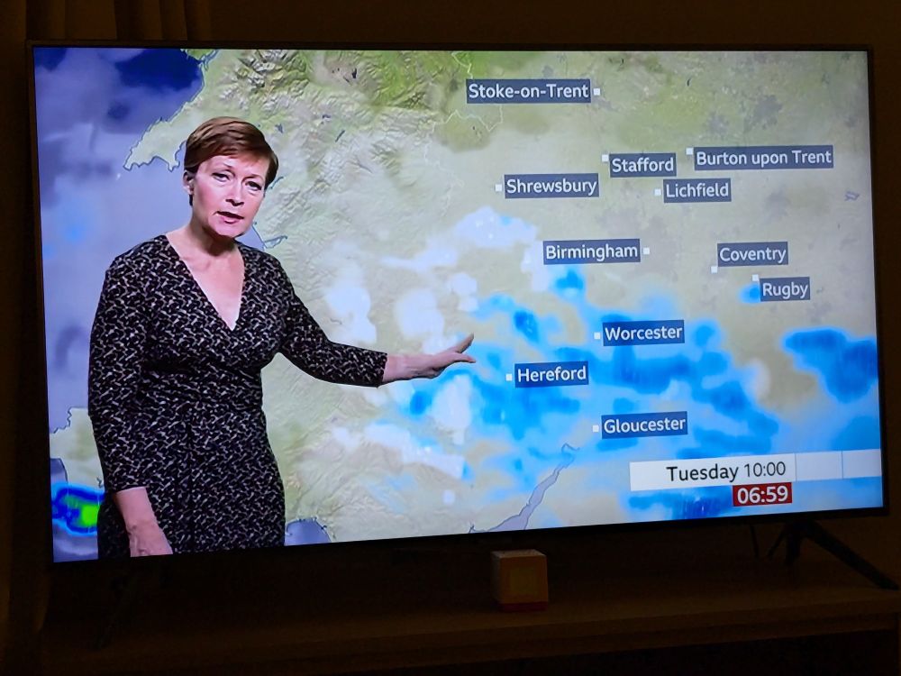 A television screen. This shows a weather forecast for the Midlands of England, highlighting Stoke-on-Trent, Stafford, Lichfield, Shrewsbury, Hereford, Worcester, Birmingham, Coventry, Gloucester, Rugby, and Burton upon Trent. The forecaster is highlighting an area of snow and rain while looking at the camera.