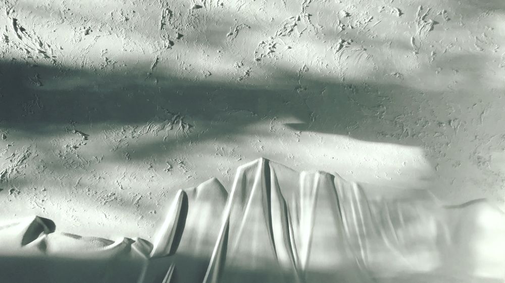 Tree shadows fall across a white wall sculpture of a soft folding mountain with texture in the sky that resembles clouds

