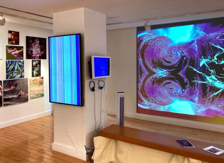 A gallery with still clusters of pictures in the background a three screen based images in the foreground. One vertical with blue green stripes. A small horizontal blue screen with headphones and a large abstract purple and white screen projection behind the column that the smaller screens hang on.