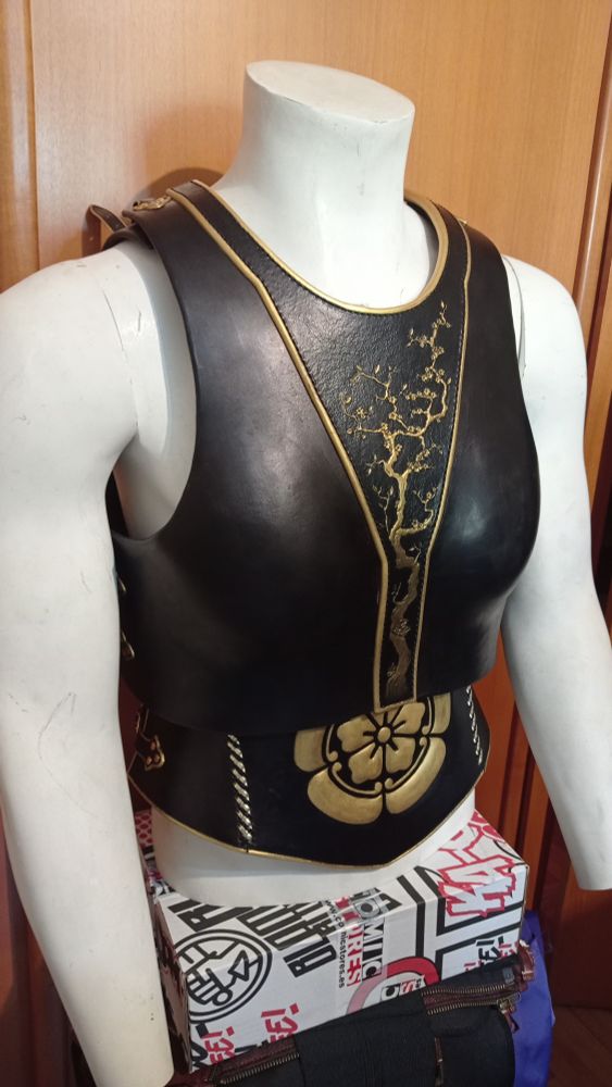 Pic front slightly towards right side of a black leather cuirass and corset. Both moulded for a female body type, thin waist, prominent hips and breasts, even though it's put on a male mannequin. Japanese inspired decorations tooled and embossed at the front (a quince branch on the cuirass, Nobunaga's emblem quince flower on the corset) and painted in golden