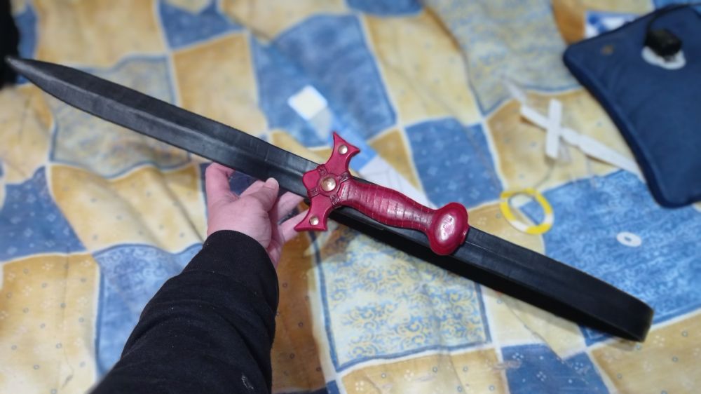 Pic of a black leather strap, slightly bent on the middle through all its length to make it look like the blade of a sword. In the center, the hilt piece carved in leather and dyed in red to see how it will look being used