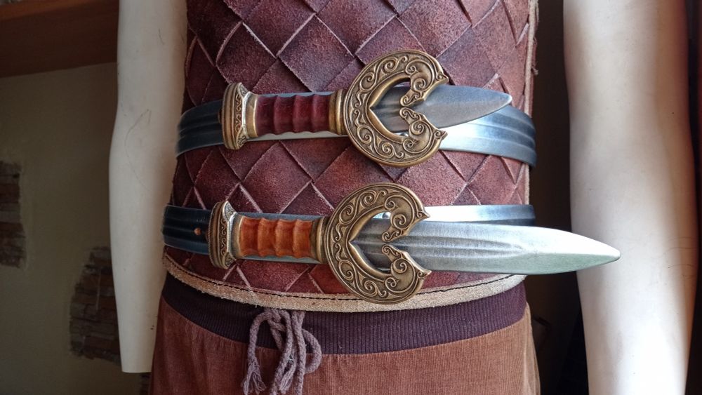 Pic of a mannequin in a leather norse-style cuirass, wearing two leather belts that looks like Thèoden sword, the aged gold and brown wood looking (they are a carved thick of leather layers) as the buckles and the silver shaped blades as straps