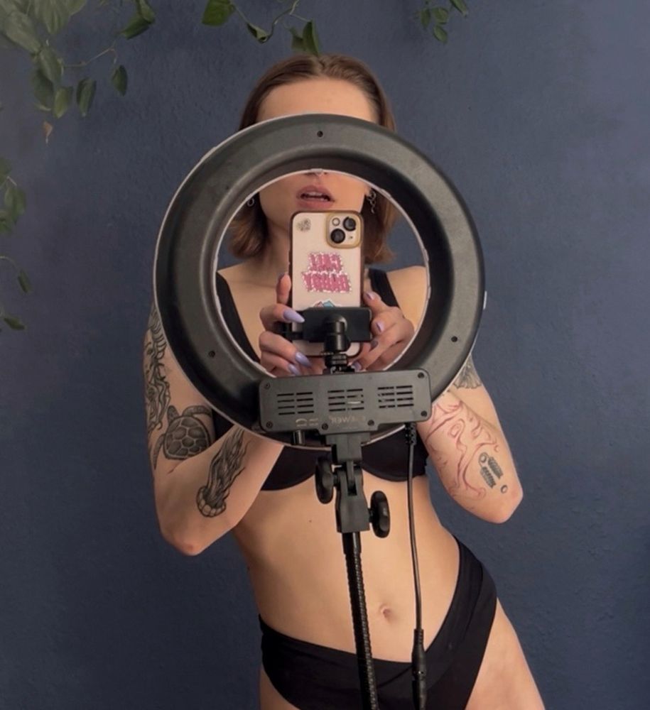 Lyra stands in front of a dark blue wall, some plants are dangling to their left side. They are wearing a black bikini and taking a photo behind the scenes. The ring light covers most of their face, only their half opened lips are not conceiled. 