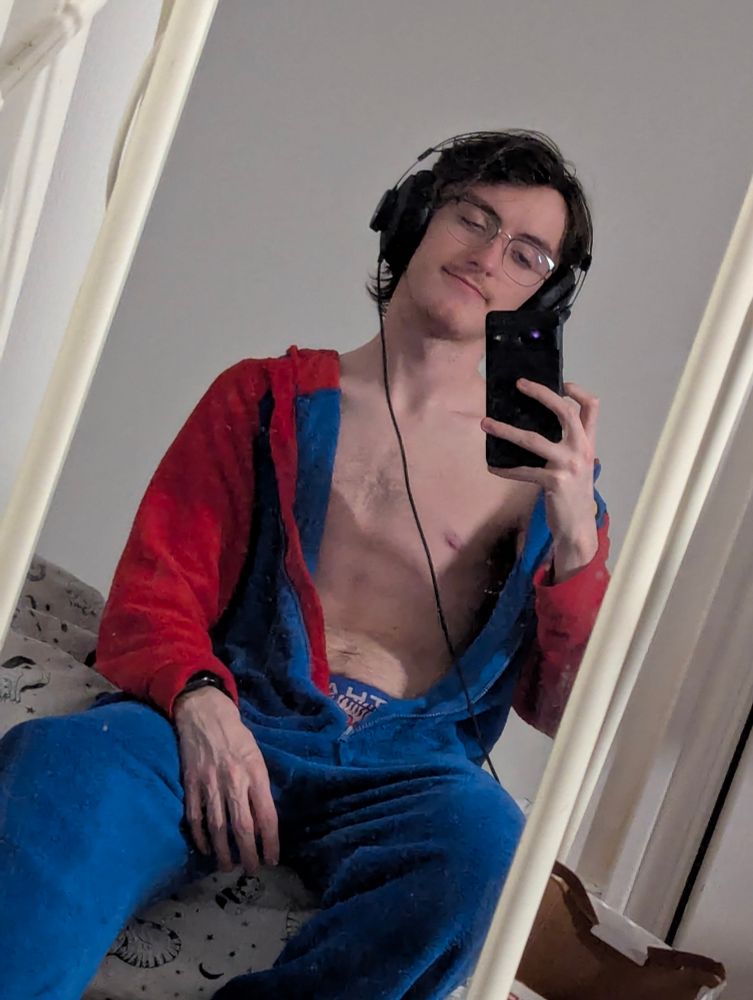 A mirror selfie of me in a Mario onesie, unzipped down to my crotch. My body is slim and and hairless except for a small tuft of light chest hair, and my face is too except for a light tache. I'm also wearing thin metal glasses and wired headphones.