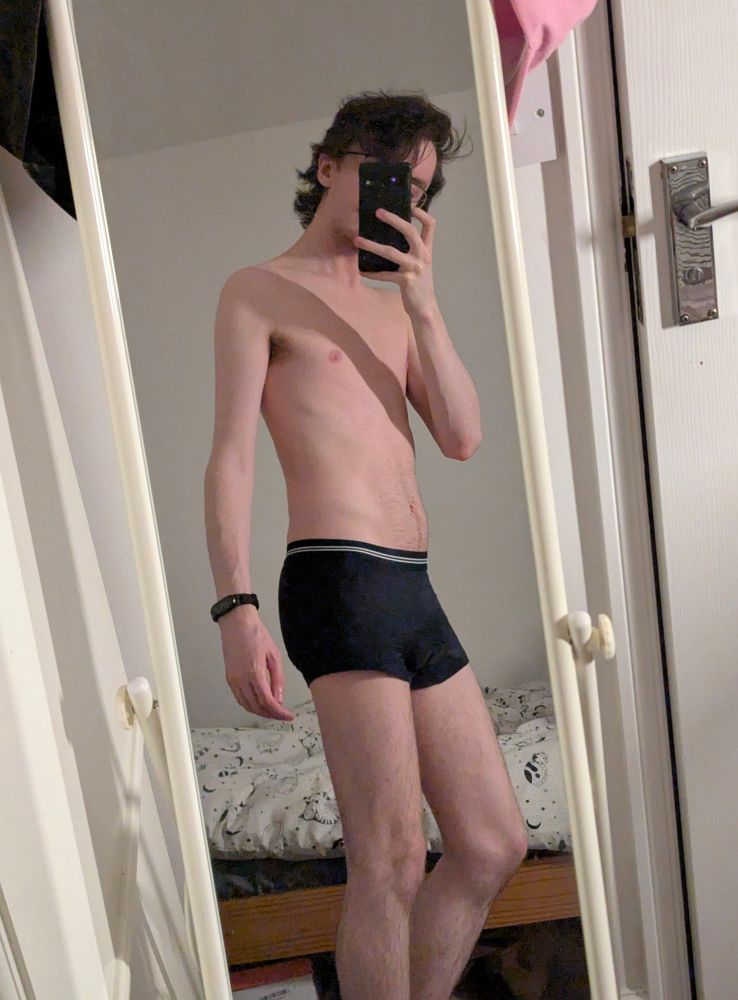 A full-body mirror selfie of me wearing only navy blue boxer briefs, which have a couple of white lines on the waistband. My body is slim and hairless, only light ginger hair on my legs. My brunette hair is thick and wavy.