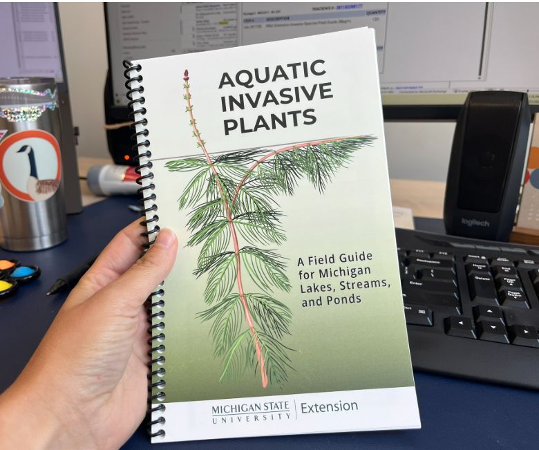 A hand holds a spiral-bound booklet entitled, "Aquatic invasive plants: A field guide for Michigan lakes, streams, and ponds", published by Michigan State University Extension. An illustration of Eurasian watermilfoil decorates the cover.