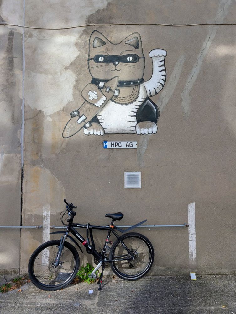 Auf einer Betonmauer eine Winke-Katze in Schwarz Weiß.
Darunter ein Fahrrad.