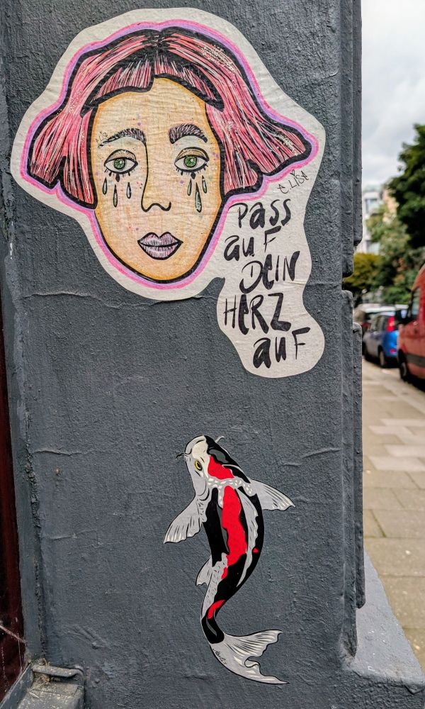 Auf einer grauen Wand ein Fisch mit rotem Rücken. Darüber ein Frauenkopf mit rotem Haar. Die Dame hat Tränen unter den Augen. Daneben das Wording; Padd auf dein Herz auf. 