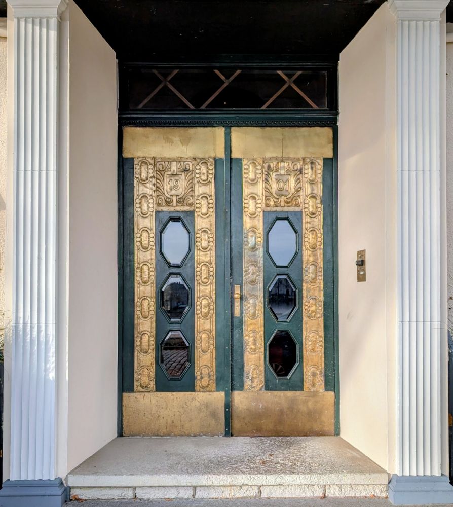 Ein kunstvoll gestalteter Eingangsbereich mit einer doppelflügeligen Tür. Die Tür besteht aus dunklem Holz oder Metall und ist reich mit goldfarbenen, ornamental verzierten Paneelen geschmückt. In der Mitte jedes Türflügels befinden sich zwei achteckige Fenstereinsätze aus Glas.

Oberhalb der Tür sind die Zahlen 19 auf der linken und 01 auf der rechten Seite zu sehen, was vermutlich in auf das Baujahr 1901 hinweist. Der Türrahmen ist dunkel abgesetzt, und über der Tür befindet sich ein schmales Oberlicht mit einem Ziergitter in X-Form.

Links und rechts rahmen zwei weiße, kannelierte Säulen den Eingang ein, die an klassische Architektur erinnern. Der Bodenbereich besteht aus hellem Stein und führt über zwei Stufen zur Tür hinauf.

Hotel Niederländischer Hof 
Schwerin
