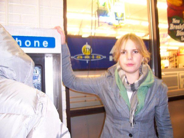 moody emo 15 year old thea making an early 2000's attempt at duck-face with heavy eyeliner, curtain bangs, a green hoodie, scarf, & blazer standing next to a phone booth.