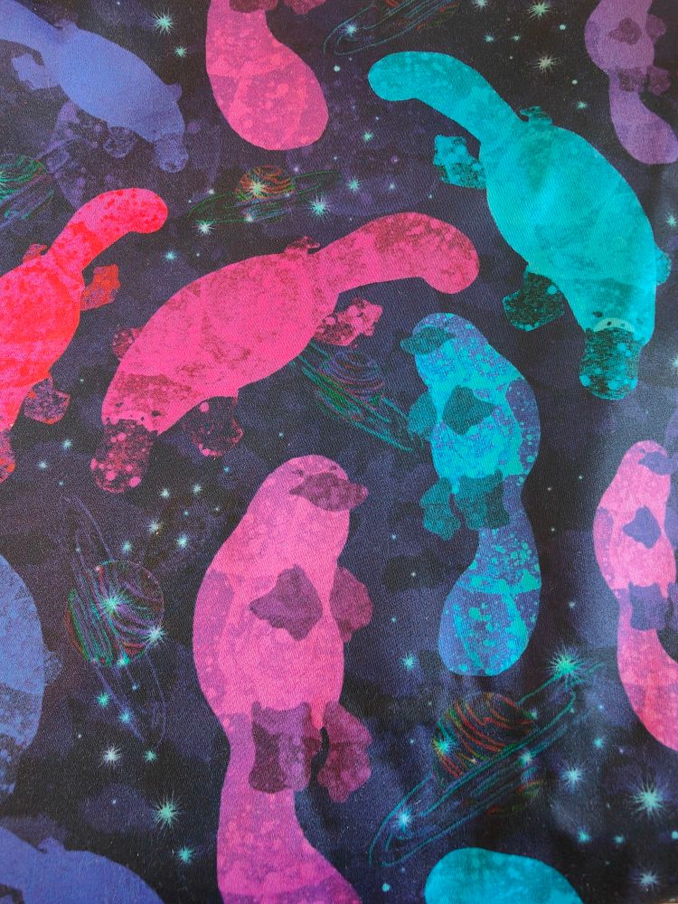 Sample of fabric with pink, purple, teal and red cartoon platypi swimming through a galaxy printed on it.