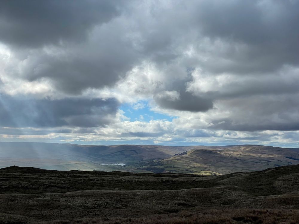 Wensleydale viewed from high on Ellerkin Scar. Shadow in the foreground. Patches of sun illuminate distant green, undulating terrain. Semerwater in the distance. Clouds with patches of blue sky allowing shafts of light to penetrate.