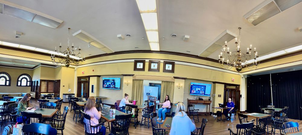 Meeting a Centre college, large room, with tables and chairs, and wonky wide angle panoramic shot that stretches everyone out