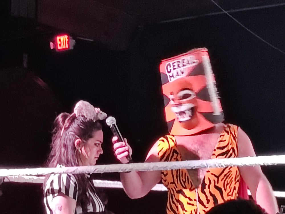 The wrestler Cereal Man holds a microphone to a rat perched on the head of a referee