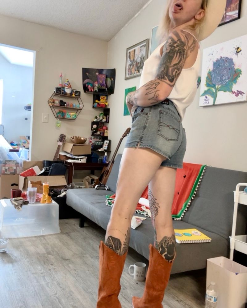 Sinéad standing in her living room in brown cowgirl boots, daisy dukes and white pencil string top and white Stetson, she’s looking over her shoulder with her tongue out. Ass looks like the peach emoji