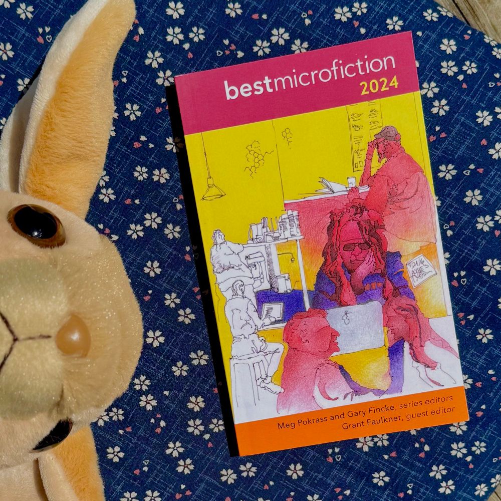 Image shows a copy of the book “Best Microfiction 2024” by Pelekinetic publishing laying on a blue floral background. Next to the book is a stuffed toy jerboa, cropped so only the head is showing, peeking out from the left side of the frame. 