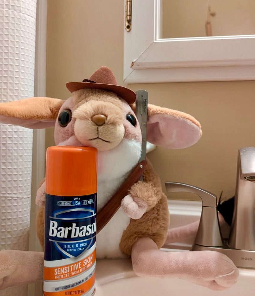 Jerbie, a stuffed jerboa wearing a brown cowboy hat, holds a straight razor while sitting on a bathroom sink behind a can of shaving cream.