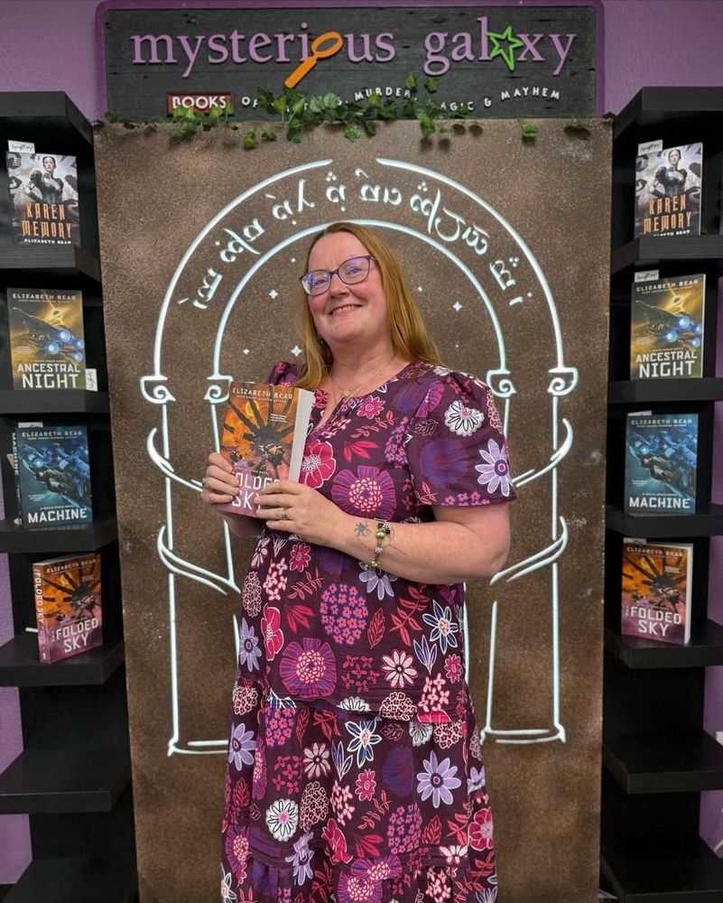 Elizabeth Bear, a white woman in a magenta dress, standing in front of the Doors of Durin under a sign that reads Mysterious Galaxy, holding a copy of The Folded Sky.

The doors are flanked by other Elizabeth Bear books. 