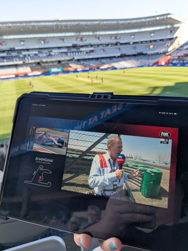 A hand with teal nail polish holding an iPad showing Formula 1 Chinese GP Free Practice 1. A live game of cricket in a stadium is visible in the background.
