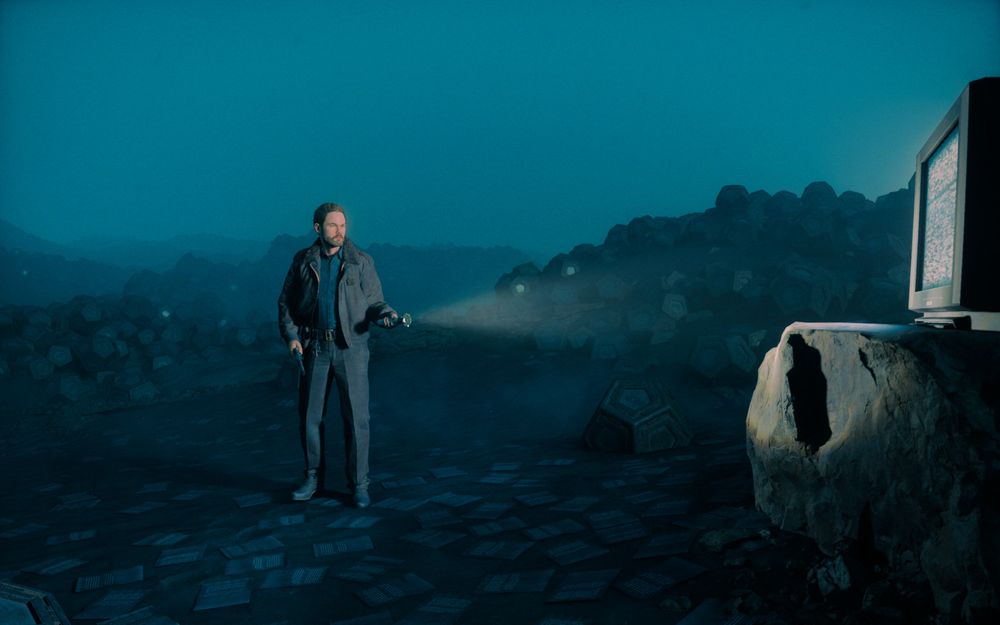 Screenshot showing 'the Actor', Shawn Ashmore, at the edge of a desolate, dark dune world. Shawn is wearing a sheriff clothes and holding a torch that is pointed at a large rock. On the rock, a TV with a static white noise sits. Behind Shawn are lots of dodecahedrons, scattered around the night desert. They seem made out of metal and some have glassy, but dirty parts that shine at the camera. Under Shawn are lots of black loose papers with white writing on them. The mood of the scene is a dark, blue, night.
