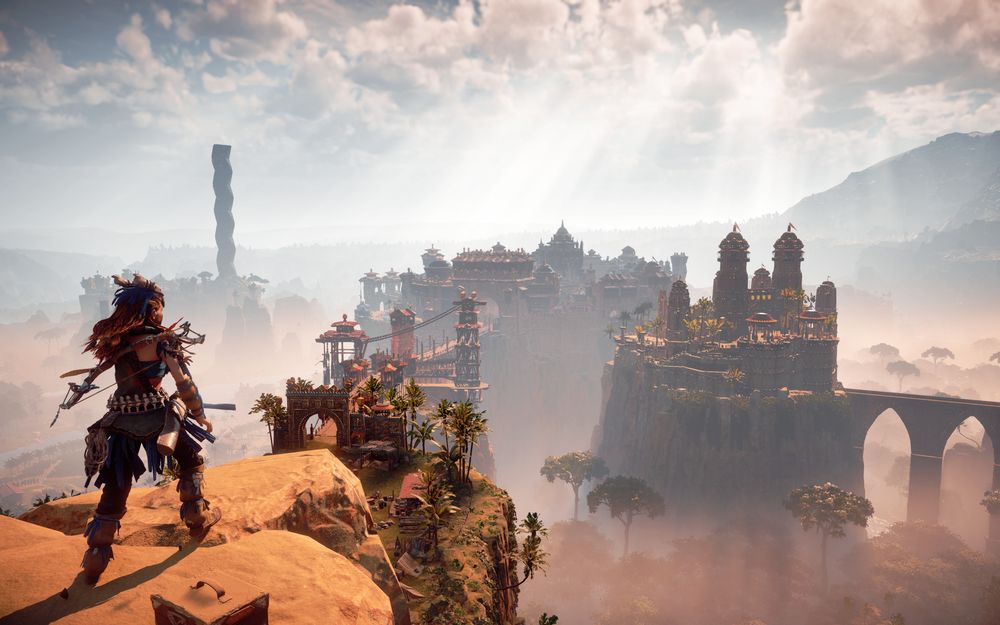 A screenshot inside Horizon, taken from a high vantage point. On the left there is the player character, Alloy. On the centre and the right side of the picture, far in the distance, among misty fog, is the city Meridian. Its stunning silhouette reveals structures made out of wood and stone. Those buildings have lots of domes. On the right side, there is a bridge made out of stone, on the left, a bridge made out of wood. It looks even larger than the stone one. The sky is bright and white clouds are scattered along. What little of the land is seen is all a brown, warm, desert. On the left side, in the far vista, stands a black obelisk with a jagged shape.