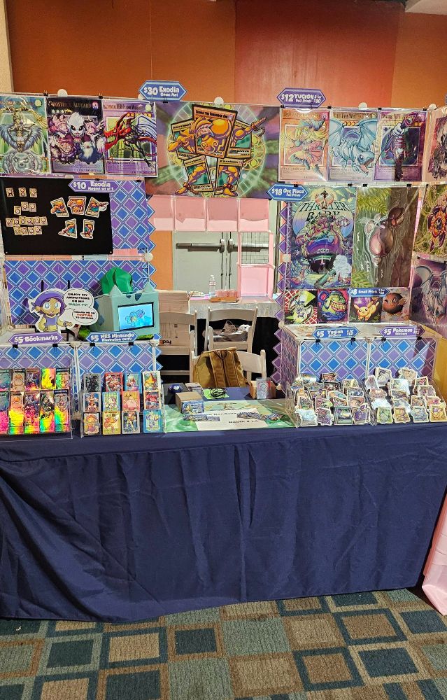 A photo of my Artist Alley table at Otakufest this past weekend. The display showcases many of my art prints and other good like stickers and bookmarks with my art on them, primarily focused on Yugioh, Pokemon and Nintendo. Also there's a small handmade TV by me thats designed to look like a Bulbasaur which plays my own animations. 