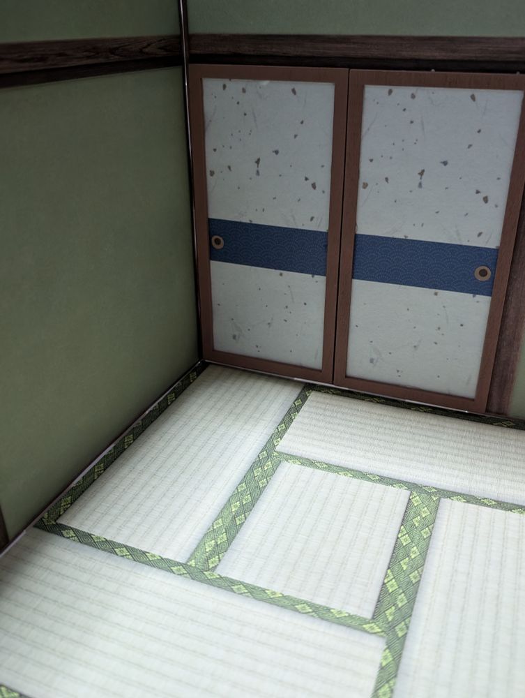ぬい用和室の写真。
畳風床に木の枠がついている風の緑の壁。
向かい正面には襖がついていて、白を基調として青い横ラインが半分より少し下に入っている。