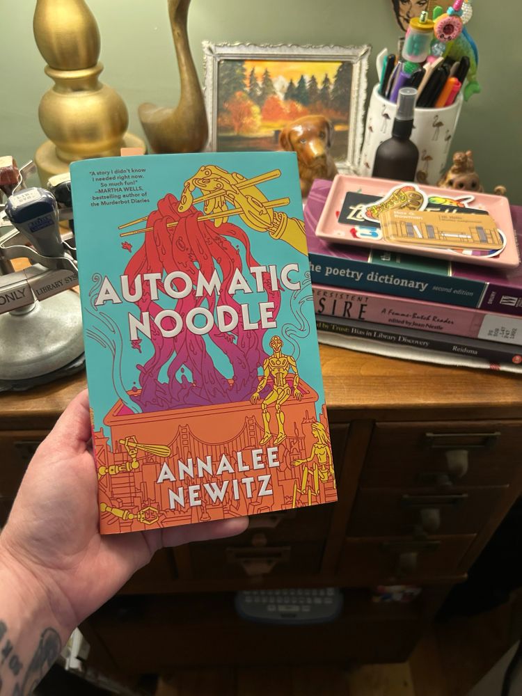 I’m holding the book, automatic noodle, by Annalee Newitz. The cover is bright, and fun — oranges, purples, red, and light blue make up the cover, and the title is in bold white letters. 