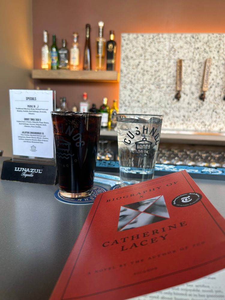 A red and black book, Biography of X, sits on a bar top in front of a glass of soda, and a glass of water; in the background there are glasses, and a full bar. 