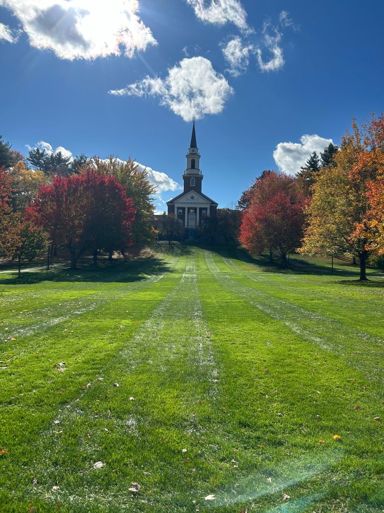 A bright green lawn leads to the view of a small chapel. The chapel is framed by trees. 