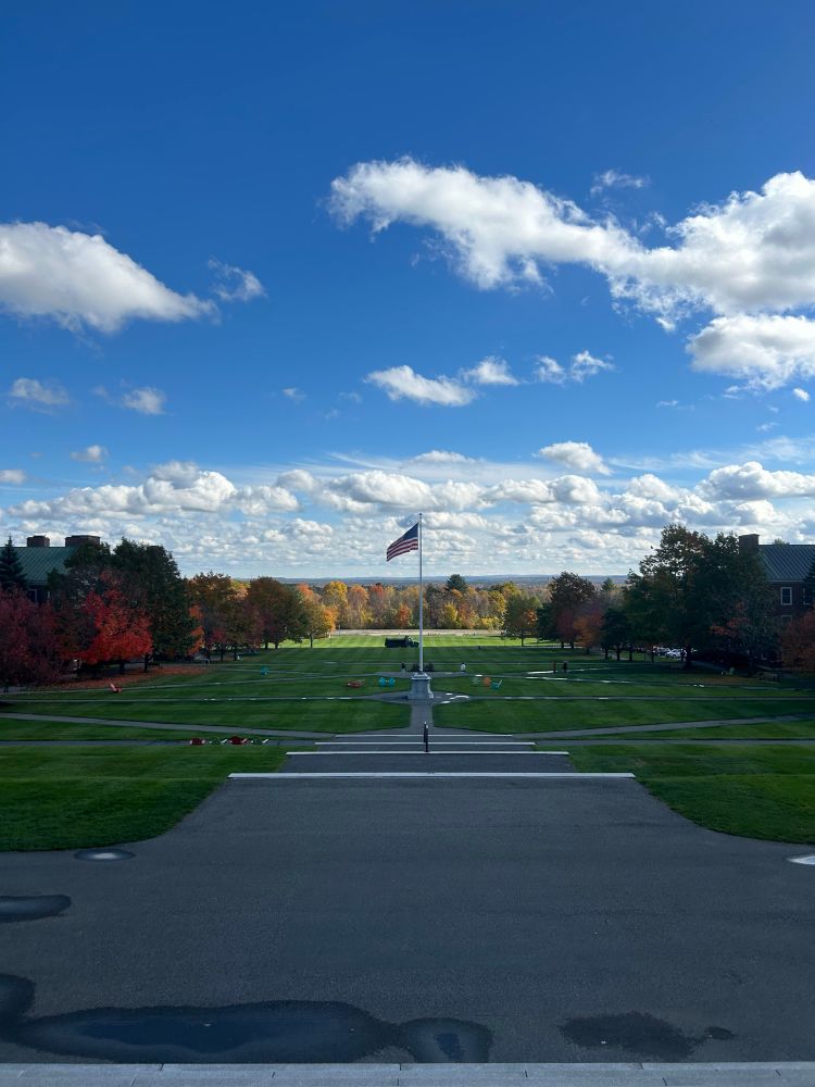 A view from the steps of Miller Library, a building that sits in the center of campus. The lawn is green, and there are trees displaying fall colors, framing each side. 
