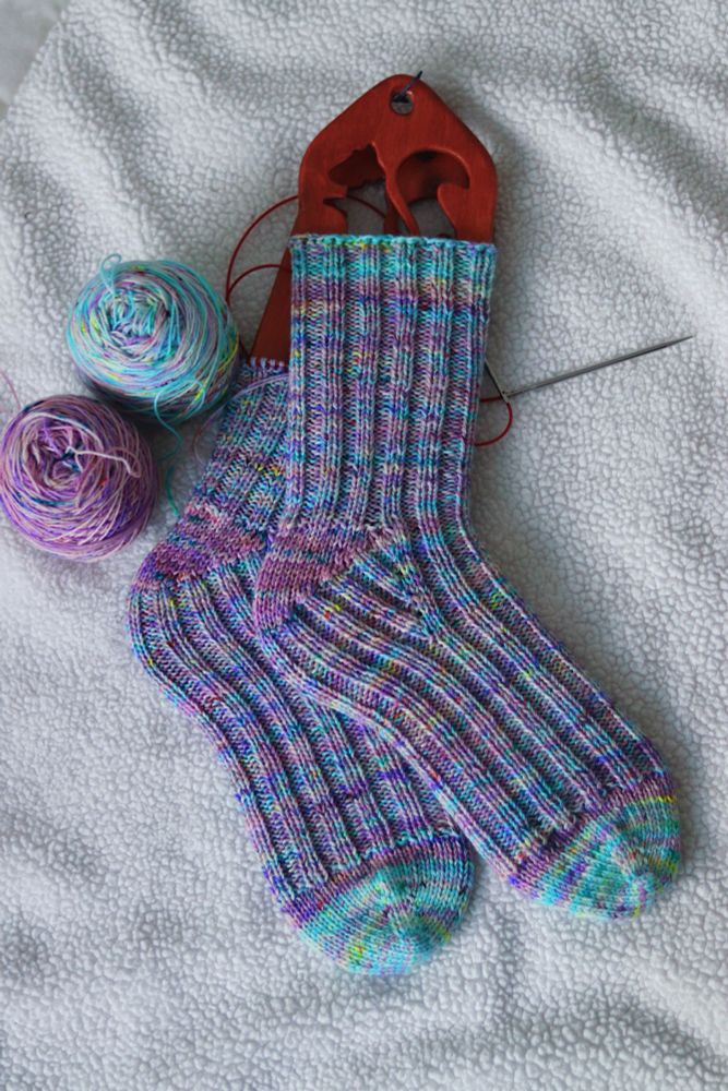 Teal and purple socks on wooden sock blockers laying on a white fuzzy blanket. 