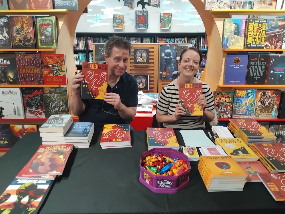 Paul and Rachael, behind a table full of books, pens and chocolates, holding up copies of Nessie. Behind them is the store's big GN/gaming books display.