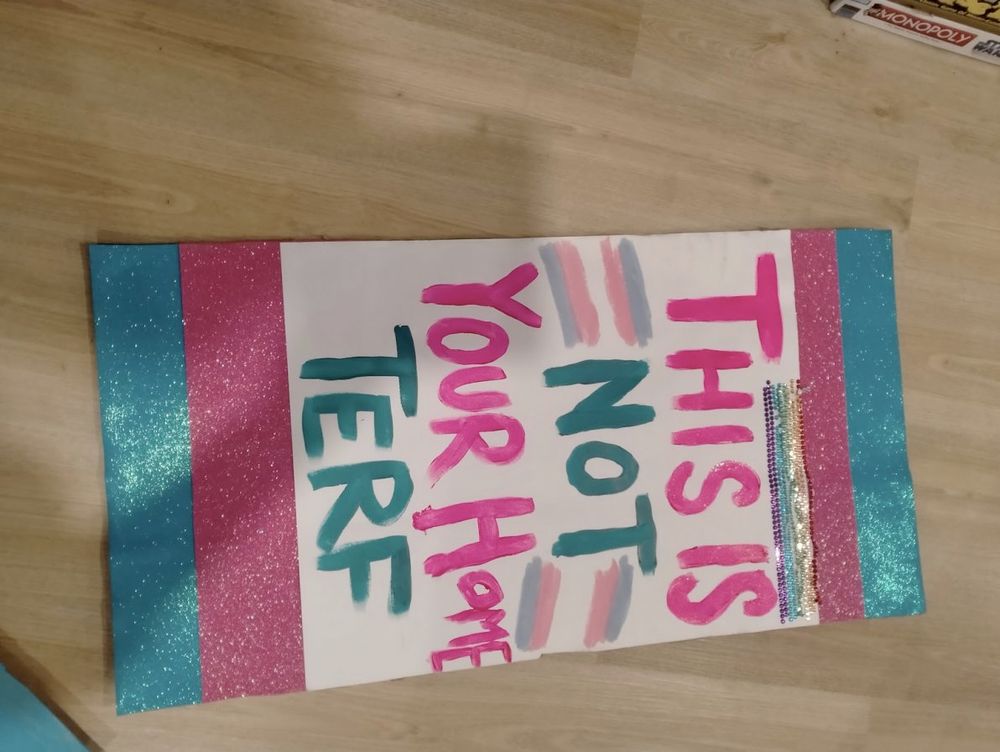 A sign made from cardboard that says THIS IS NOT YOUR HOME TERF. There are sparkly pieces of paper glued onto it so the sign forms a trans flag. There are two trans flags painted onto the section of the sign with the text, and strips of adhesive rhinestones that make a rainbow flag
