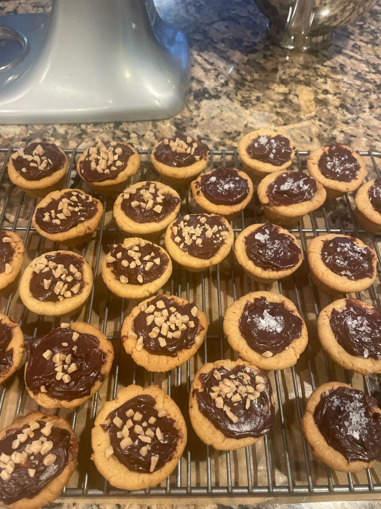 Cooling rack with many cookies on top. Cookies are tan cups (peanut butter) with chocolate filling in the center and topped with toffee or sea salt. 