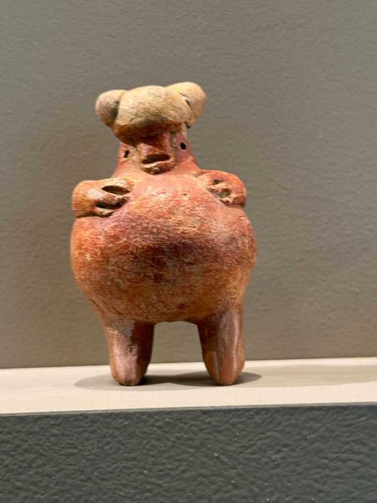 A little reddish clay figurine. It is mostly round with two legs and little arms with three fingers. The face has an opening for a mouth and two dots for eyes. The head is sort of bulbous but also has two round balls for ears I guess? I don’t know that this is—maybe a bear 